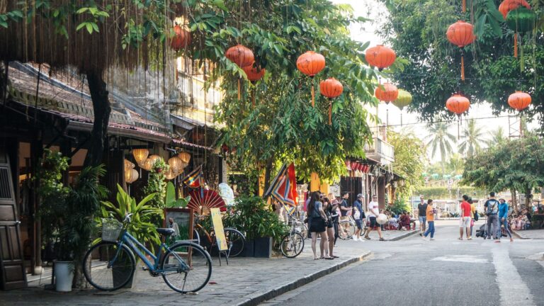 Street scene in Hoi An, Vietnam with red lanterns hanging from trees, bicycles parked by shopfronts, and pedestrians walking in warm afternoon light.