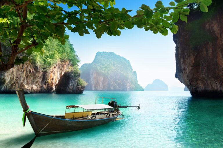 Long tail boat floating in clear turquoise water between limestone cliffs and jungle covered islands under a bright blue sky.