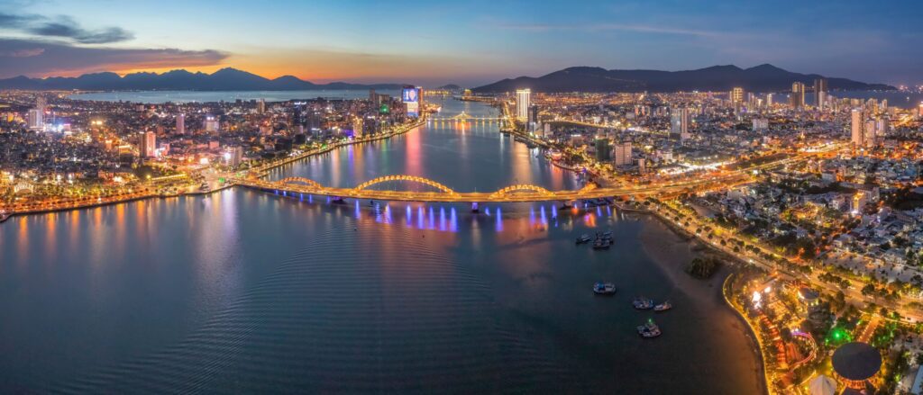 Panoramic aerial view of a coastal city at dusk, with an illuminated bridge crossing a wide river and city lights reflecting on the water.