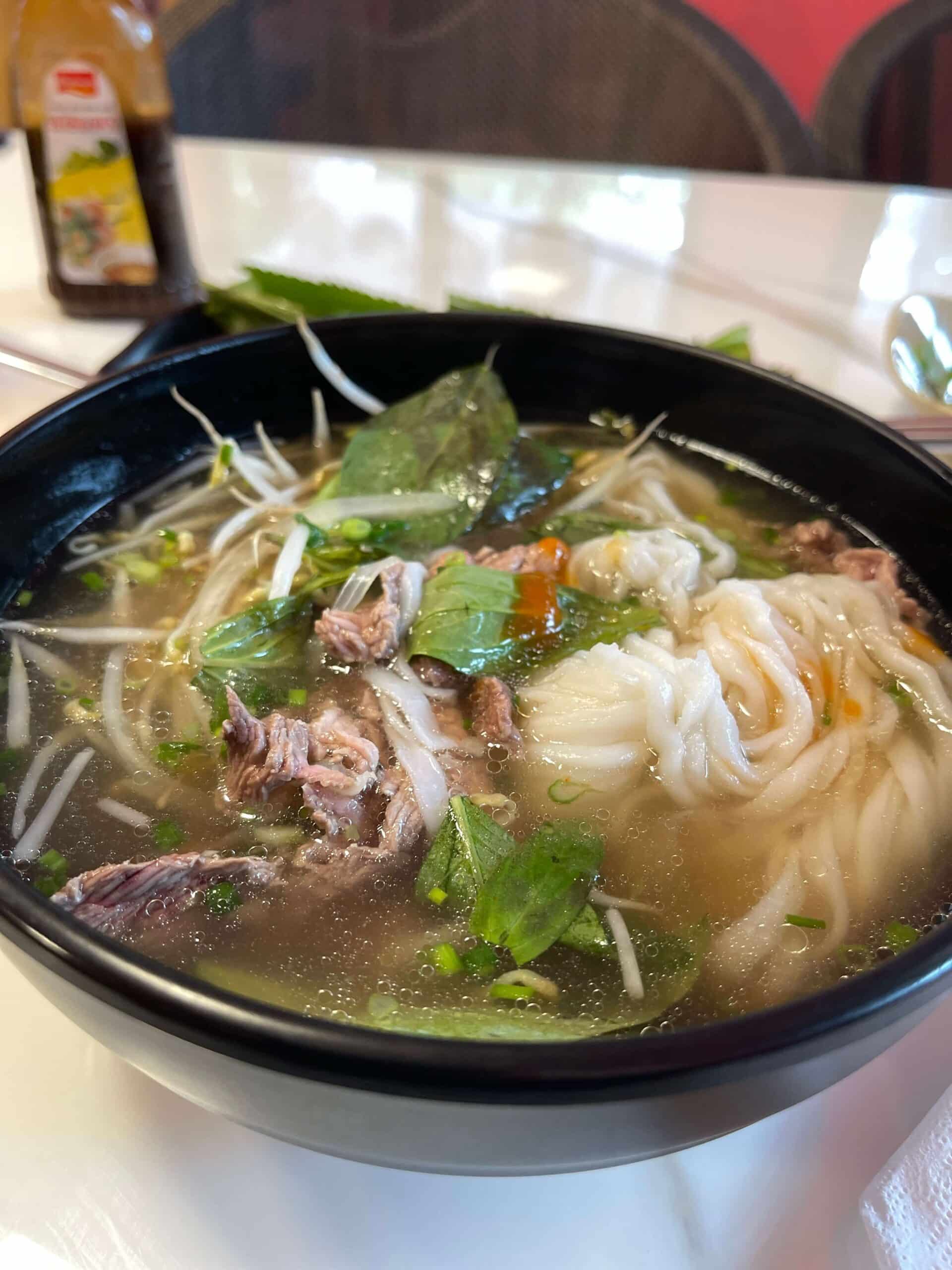 Close up of a steaming bowl of pho in Da Nang with beef slices, rice noodles, bean sprouts, Thai basil, scallions, and a slice of chili in clear broth.