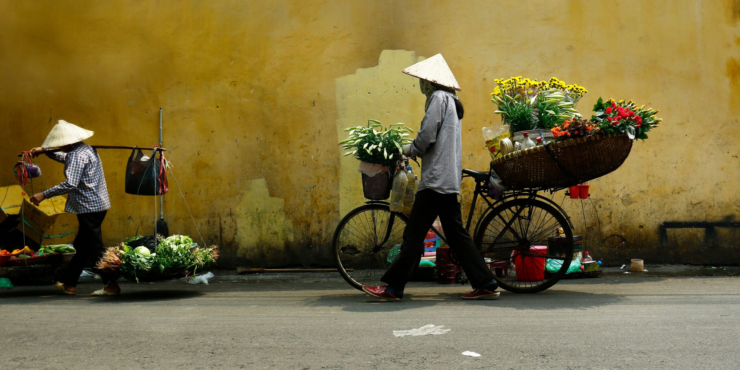 Two street vendors in conical hats pass a mustard yellow wall, one pushing a bicycle loaded with colorful flowers.