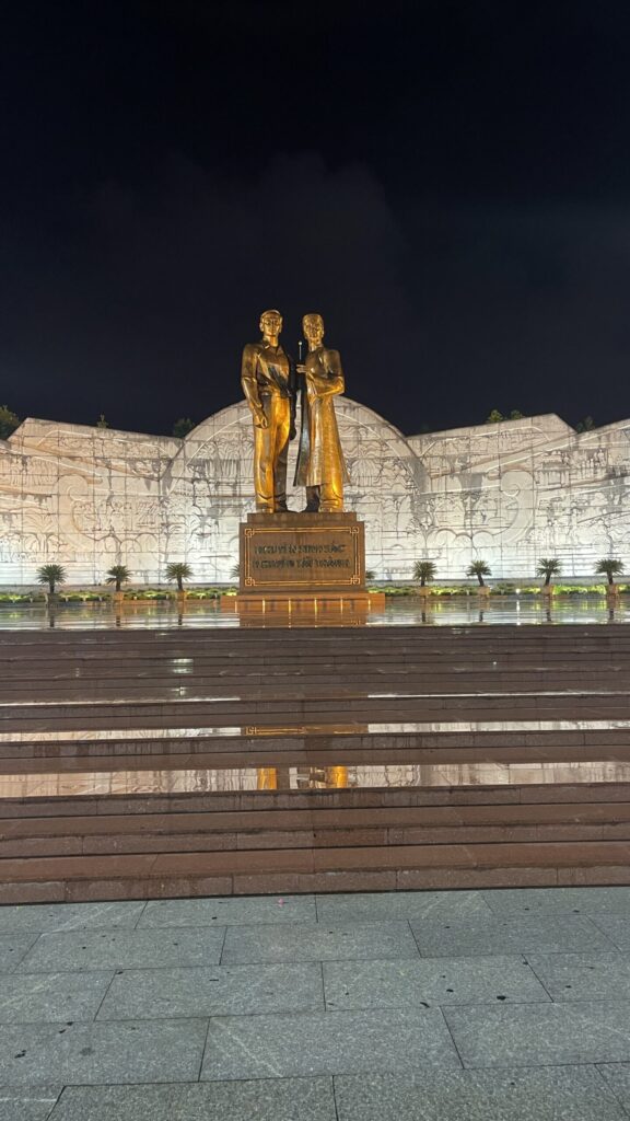 Two gold statues on a pedestal in front of a large illuminated relief wall, reflected in wet stone paving at night.