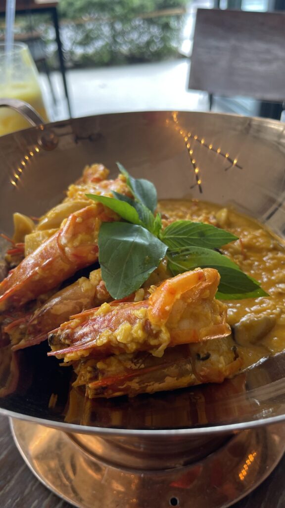 Close-up of prawns in a rich orange curry sauce, topped with fresh Thai basil in a metal bowl.