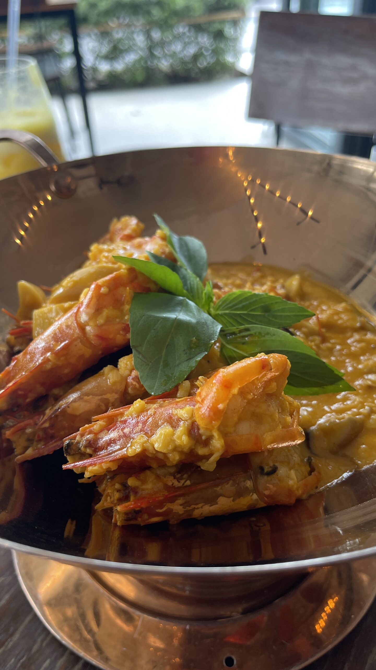 Close-up of prawns in a rich orange curry sauce, topped with fresh Thai basil in a metal bowl.