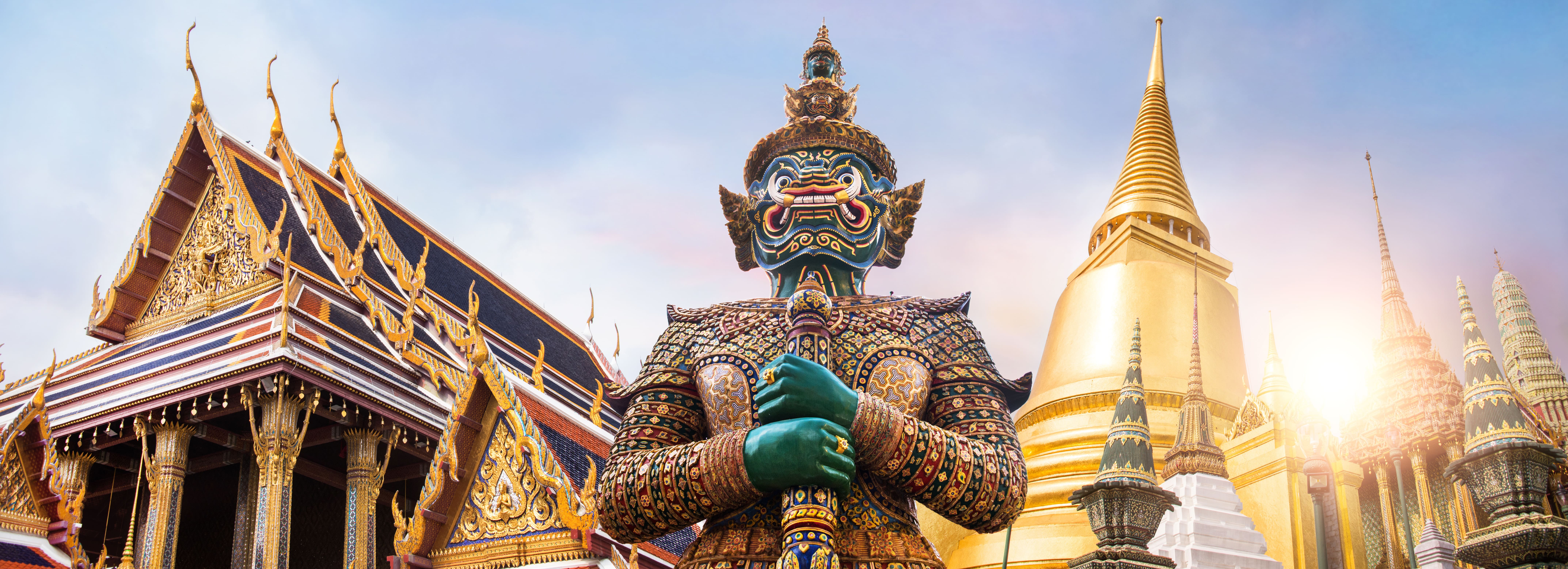 Giant guardian statue at the Grand Palace in Bangkok with ornate temple roofs and golden stupas glowing in soft morning light.