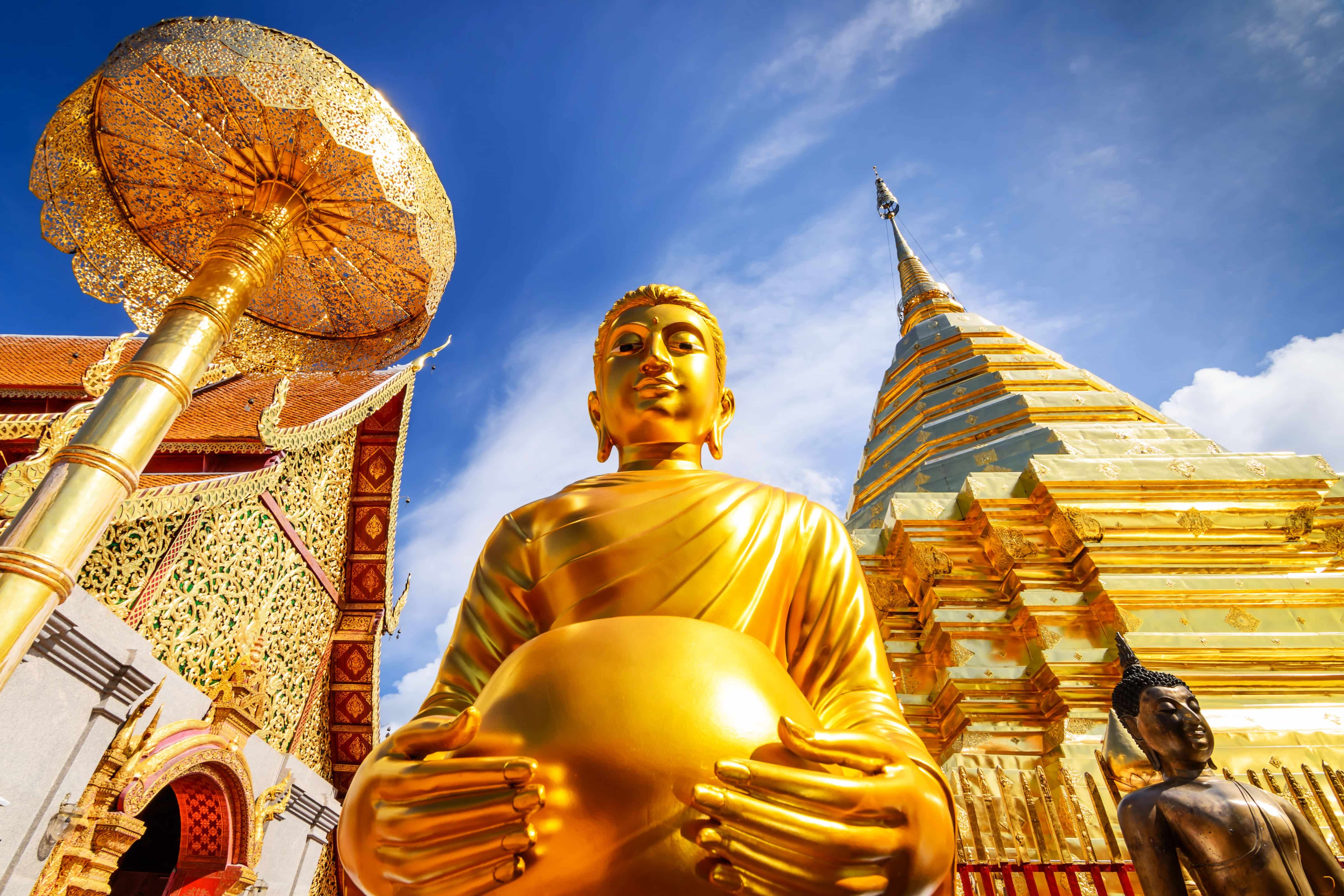 Golden standing Buddha statue holding an alms bowl beside a gold chedi and ornate temple details under a bright blue sky in Thailand.